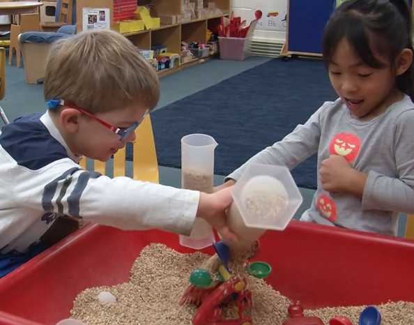 image of two young children in classroom. Boy on left is wearing glasses and pouring sand in sandbox. Girl on the right is holding cylinder tube with sand.