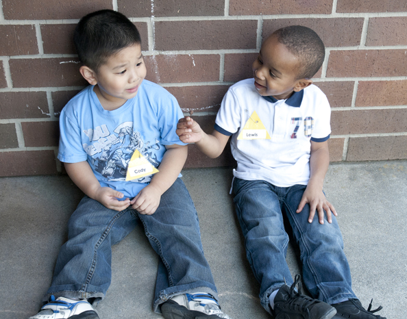 image of two young children sitting on sidewalk smiling. Boy on right is wearing white shirt and jeans. Boy on left is wearing blue shirt with nametag and jeans.