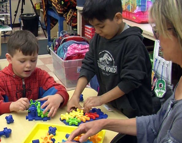 image of two young male students playing with learning blocks. Teacher on right side of image is helping students with activity. Student on left side is wearing a spiderman jacket. Student on right is wearing black Jurassic world sweatshirt.