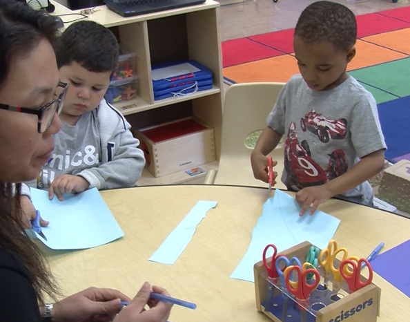 image of students and teacher in a classroom. The two young boy students are cutting blue paper with scissors. The teacher is supervising and holding a pen.